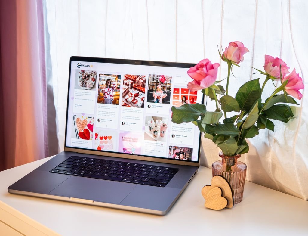 A romantic desk setup featuring pink roses in a vase next to a laptop displaying social media for Valentine's Day.