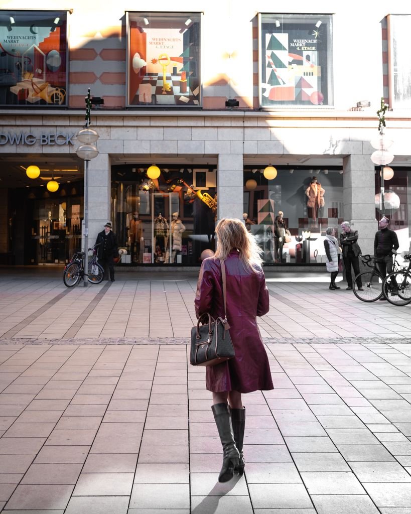 A woman in a purple coat walks past Ludwig Beck in Munich, Germany, showcasing urban fashion.