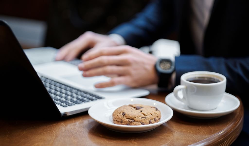 Professional working on a laptop over coffee and cookie in a cozy coffee shop setting.